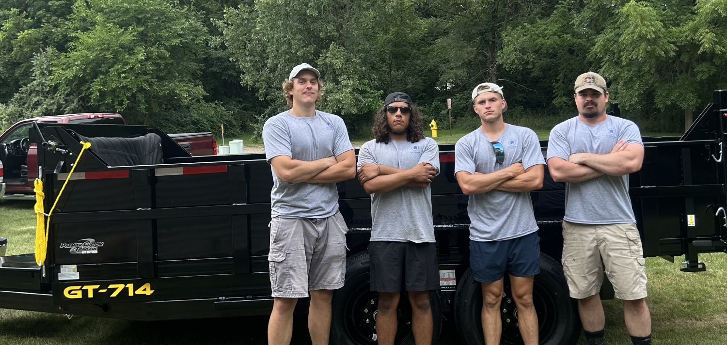 Four men in matching gray shirts standing with crossed arms in front of a black trailer