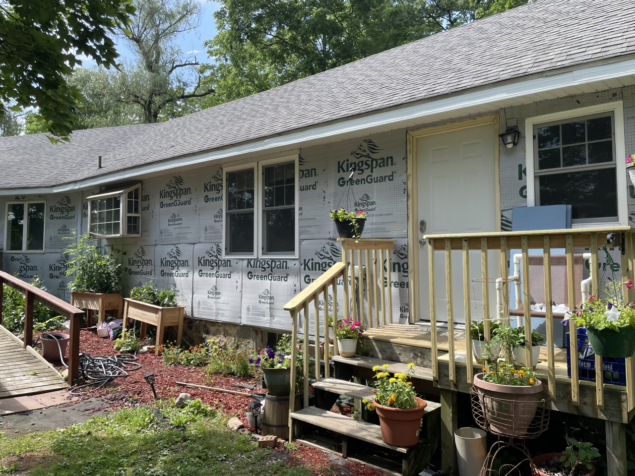 Side of a house under renovation with insulation wrap, potted plants, and wooden stairs